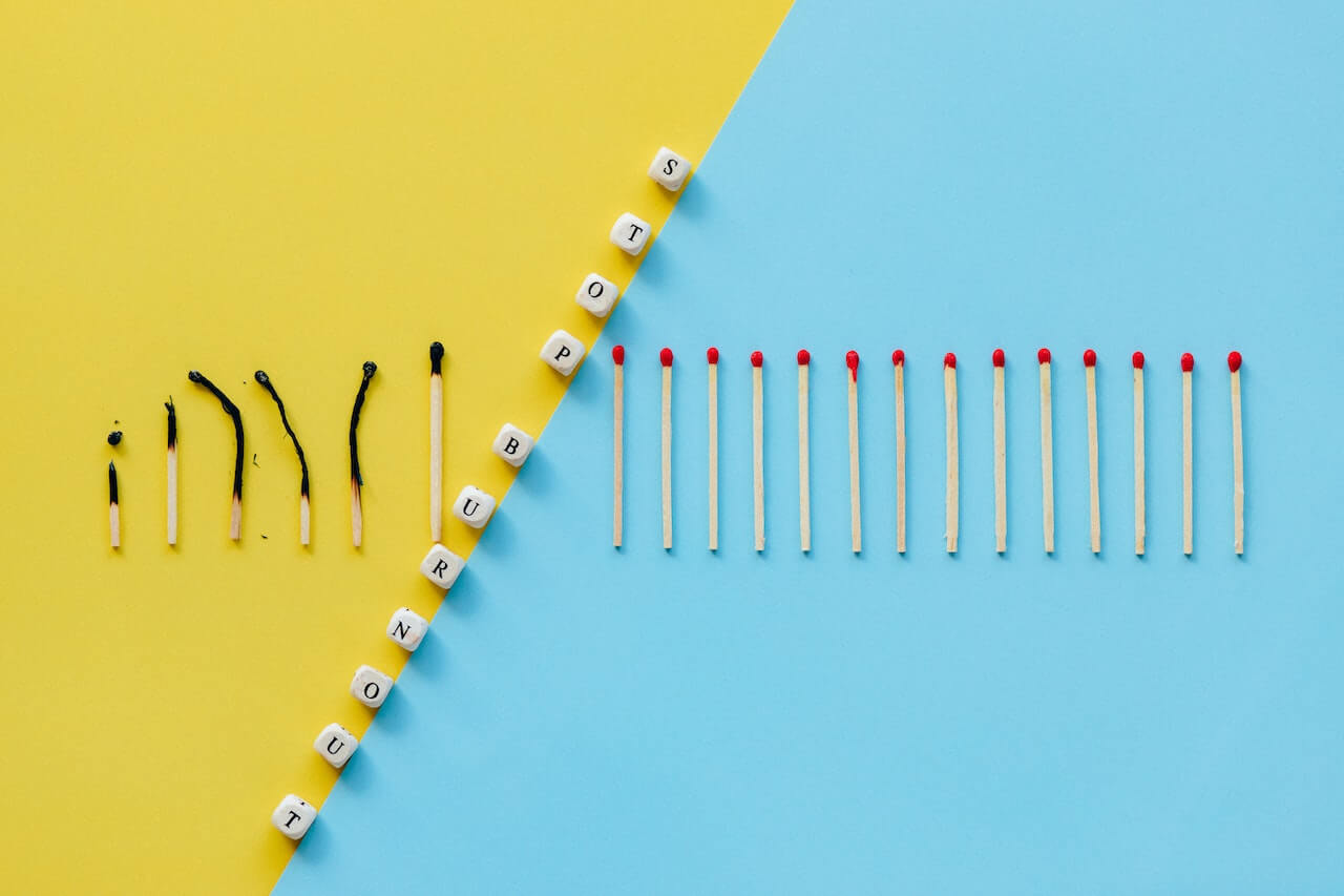 A colorful background divided diagonally by lettered dice; the background is yellow on the left side, light blue on the right. The dice spell out the words: Stop burnout. The left side has 6 burnt matchsticks next to each other. On the right side of the divide there are 14 unburnt matchsticks lined up.