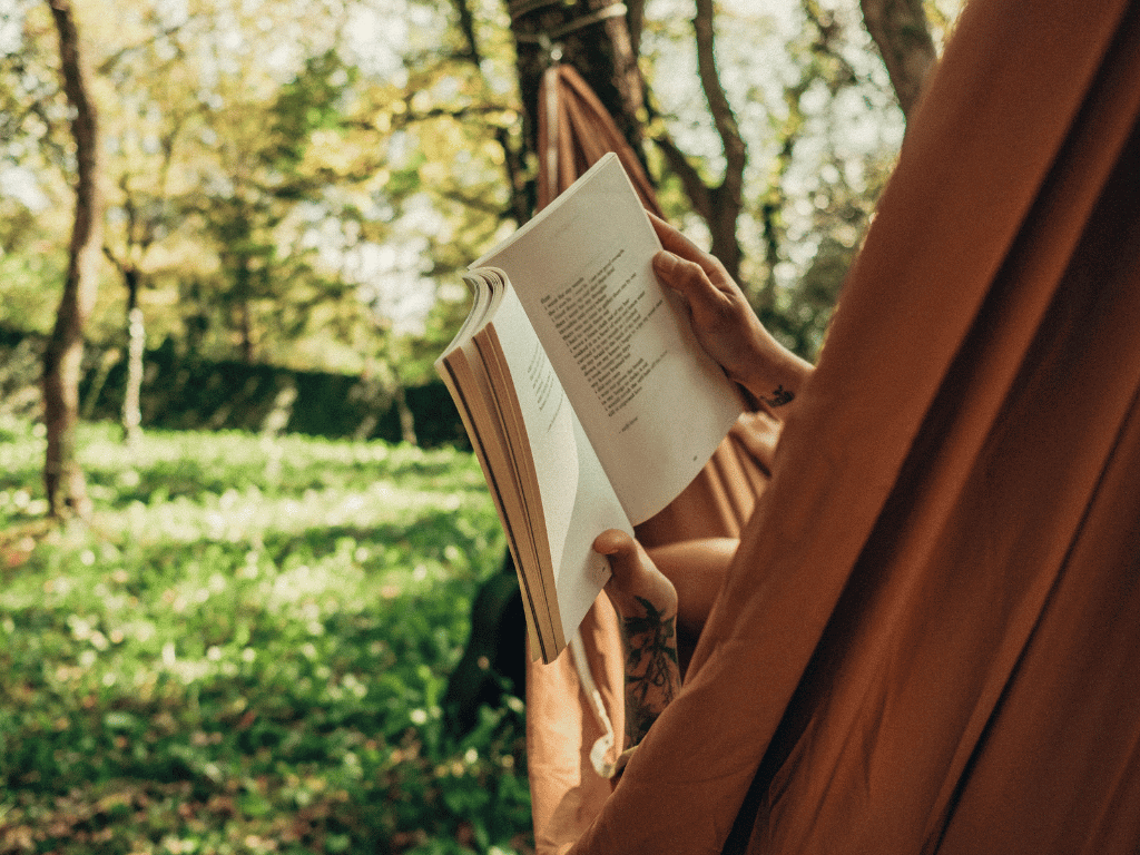 A brown hammock hanging between two trees. A person is lying in the hammock with only their tattooed arms visible that are holding up a book. In the background out of focus are green trees and grass.