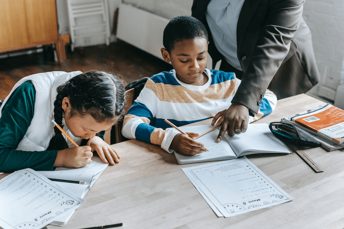 Two children sitting at a desk doing their homework. An adult is helping them with their homework and pointing at the worksheet they're explaining.