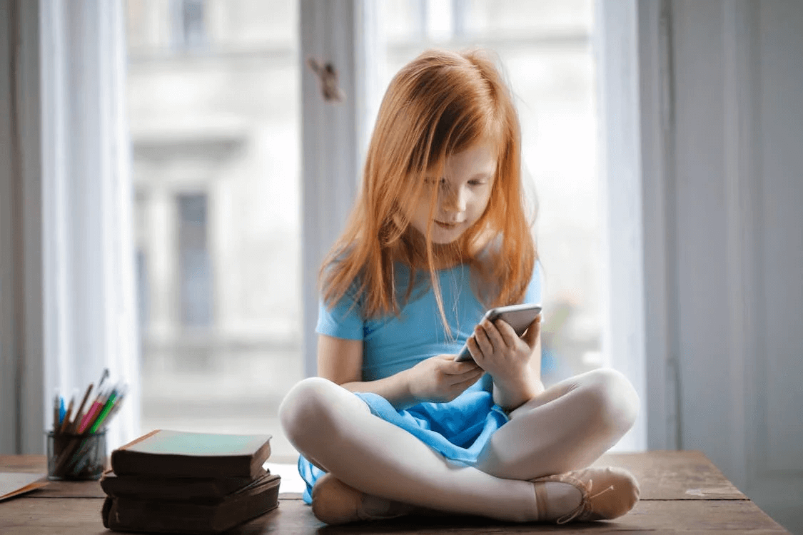 Young girl with red hair in a blue dress sitting on a table looking at a phone screen.