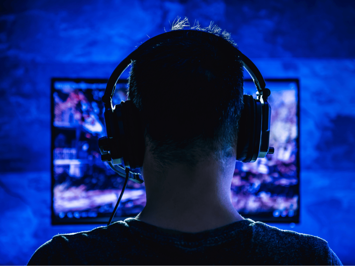 A man sits in dark room wearing a gaming headset and facing an illuminated screen.