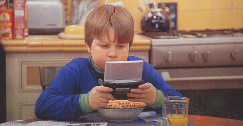 A young boy playing on a handheld device pushes his face into a bowl of cereal to take a bite so he doesn't have to let go of his game. 