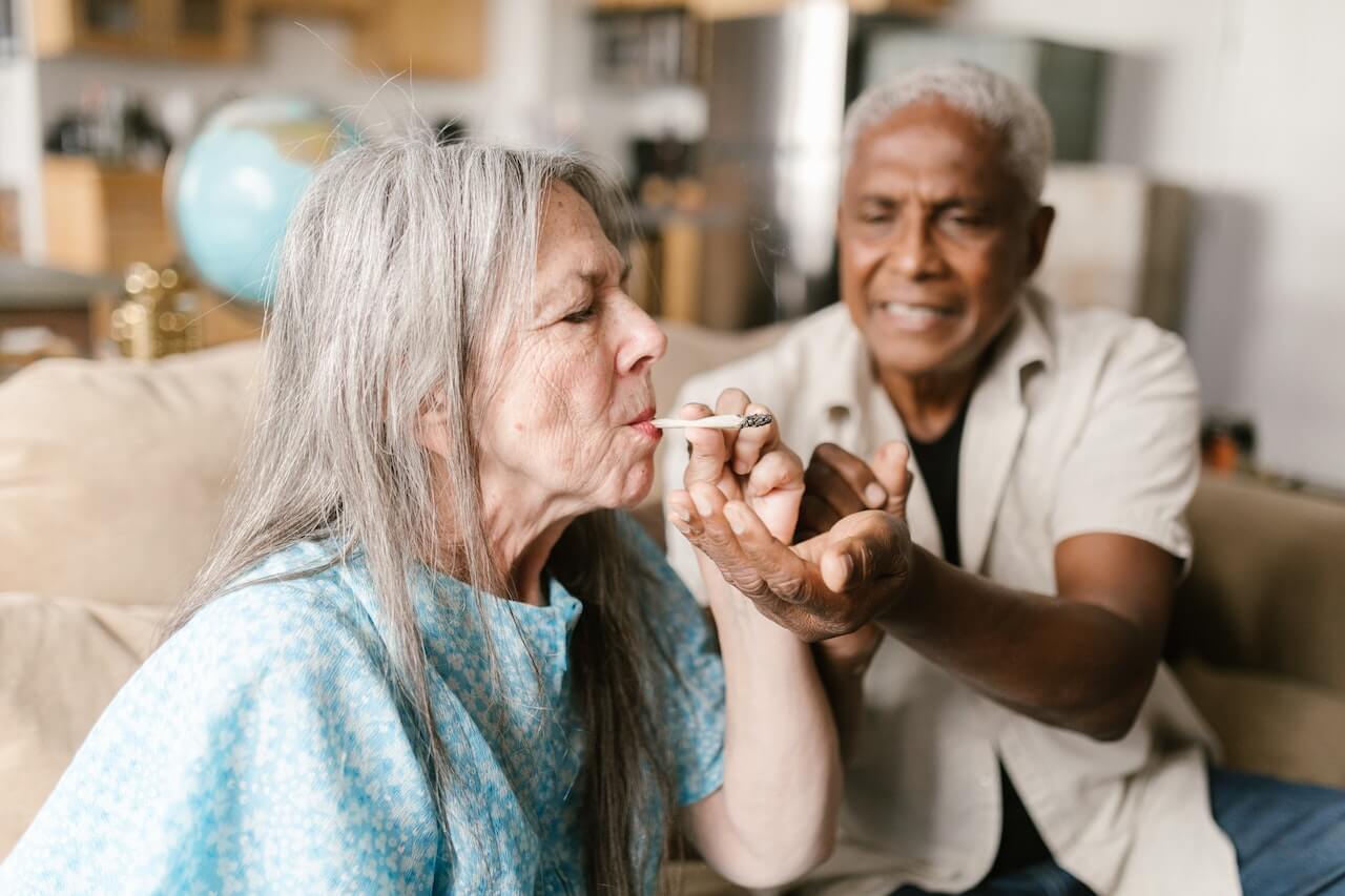 Two old people smoking a joint of cannabis or weed. An elderly black man catches the ash from the joint being smoked by an elderly white woman. 