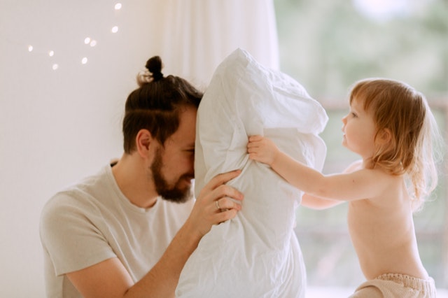 a father and young daughter holding a pillow. The dad is hiding behind it and the toddle is looking for him.