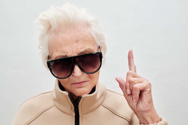 Photo of an old woman wearing sunglasses and pointing up with her index finger