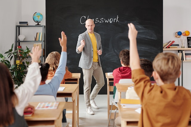 A male teacher standing in the front of a classroom with children raising their hands. He is pointing to a student.