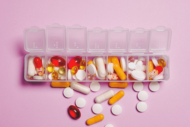 A daily pill container with a variety of medications in each slot. The background is a bright pink. The image represents object constancy and not object permanence.