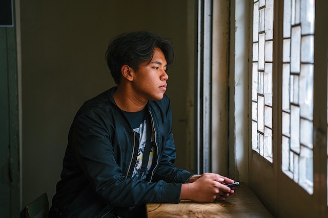 a man staring out of a window with a sad look on his face, holding a phone in his hands
