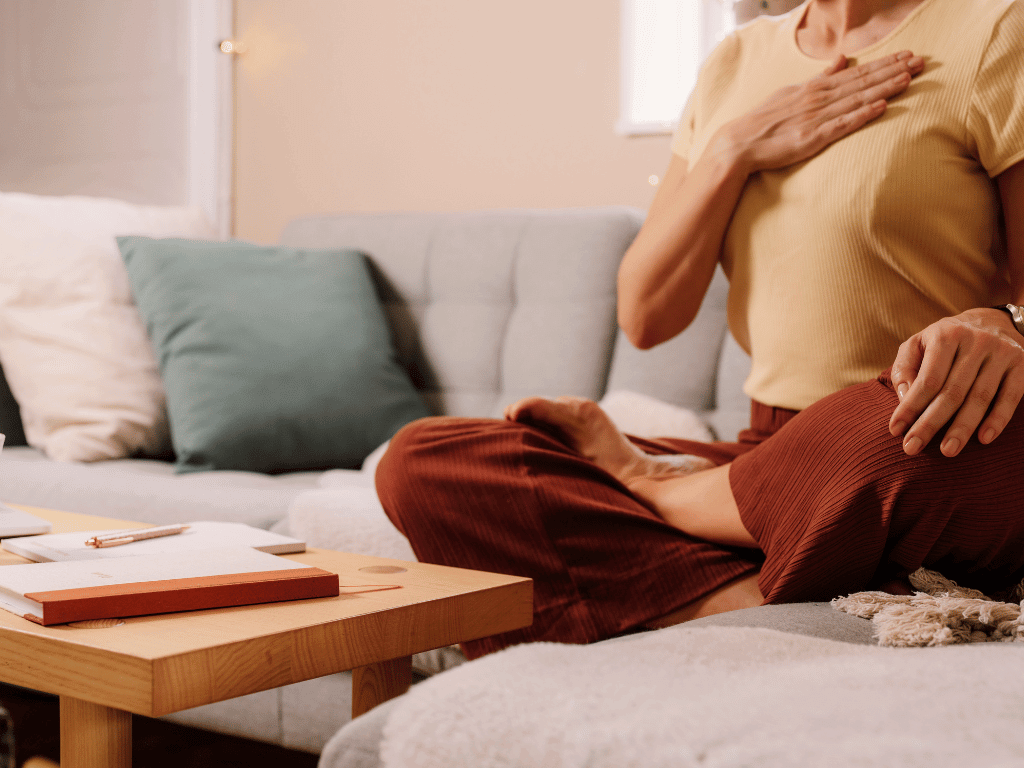 A person wearing a beige t-shirt and brown flowy trousers sitting on a grey sofa in a cross-legged or lotus position. One of their hand is resting on their left knee, the other hand is flat on their chest. There's a wooden coffee table with a notepad, a book, and a pen in front of the person. The surroundings are in light pastel colors and beige.