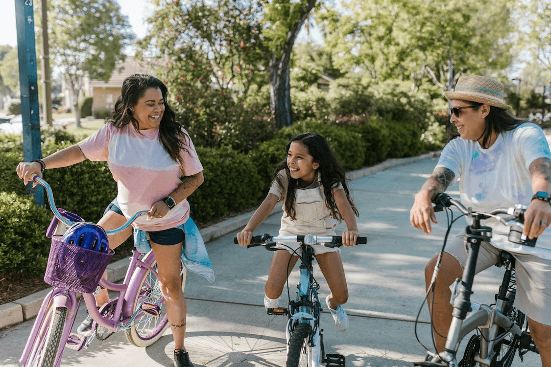  A BIPOC rainbow family of three smiling at each other while riding their bikes in a park.