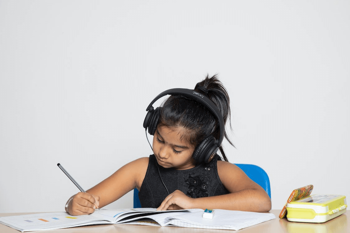A young BIPOC girl sitting at a table wearing large, over-ear headphones and writing in a notebook.