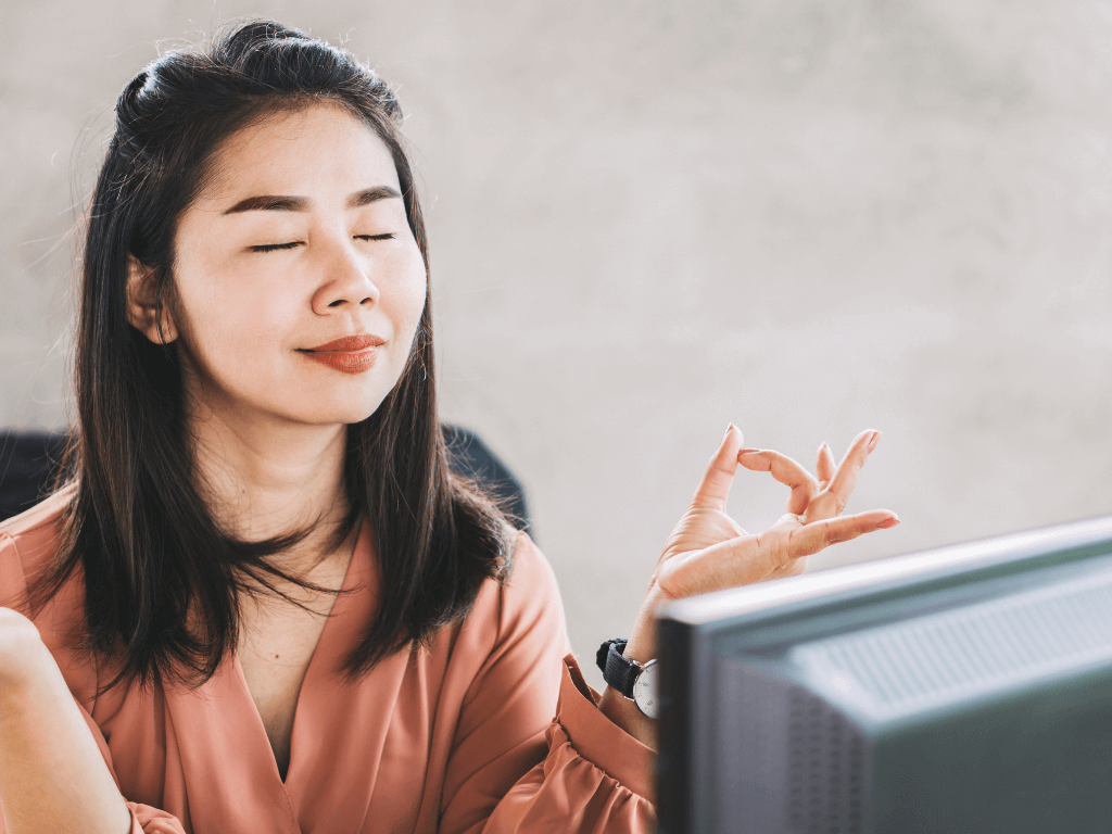 An Asian American woman calmly meditating at work to regulate her emotions.
