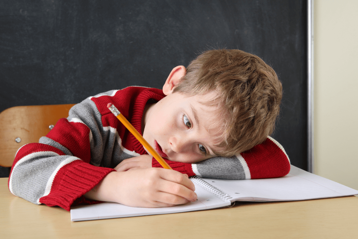 A bored child with ADHD works on his homework at school, lying with his head on his desk.