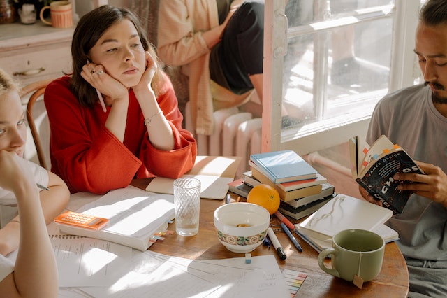 a group of three college students sitting around a table in a cafe. The young woman in red is staring off into space, bored, while the others work.