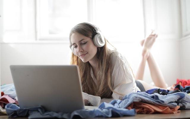 a oman with white headphones on, lying on her stomach and on her laptop. Surrounding her is a pile of clothes
